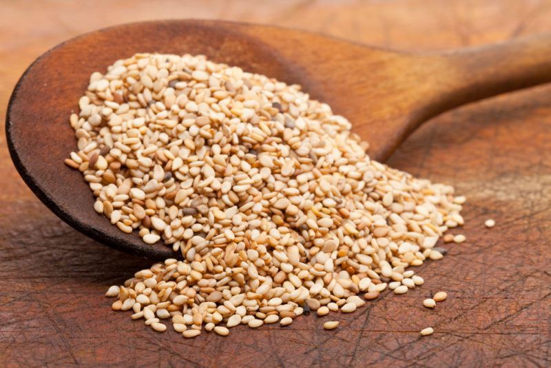 A wooden spoon filled with sesame seeds, with some spilling out onto the table