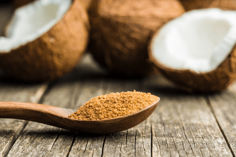 A wooden table with a spoon of coconut sugar and some coconuts in the background