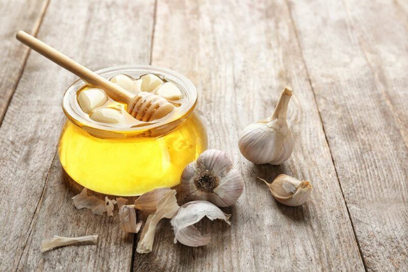 A wooden table with a small jar of honey with garlic on the top, next to some whole bulbs of garlic