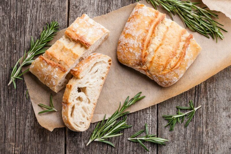 A wooden table with half a loaf of bread, plus a few slices and a variety of rosemary sprigs scattered around