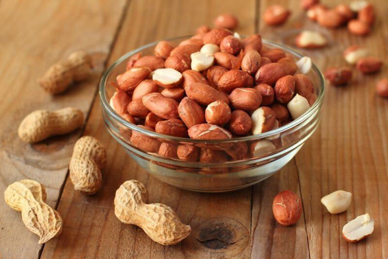 A glass bowl containing peanuts in their skins, on a wooden table with more peanuts scattered around. Some of the peanuts are still in their shells, while others are just in skins