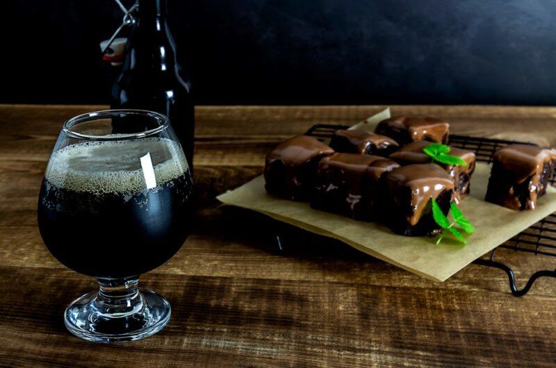 A dark wooden table with a glass of dessert stout, brownies, and a bottle