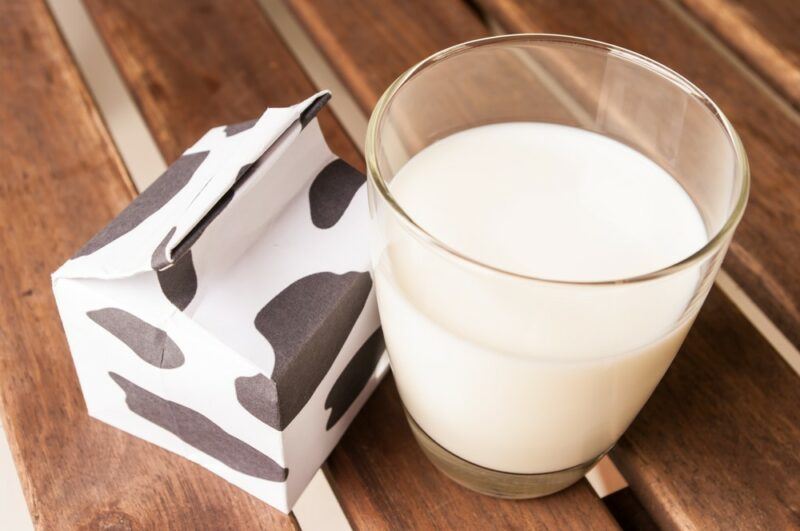 A wooden table with a glass of whole milk and a carton decorated like the spots on a cow