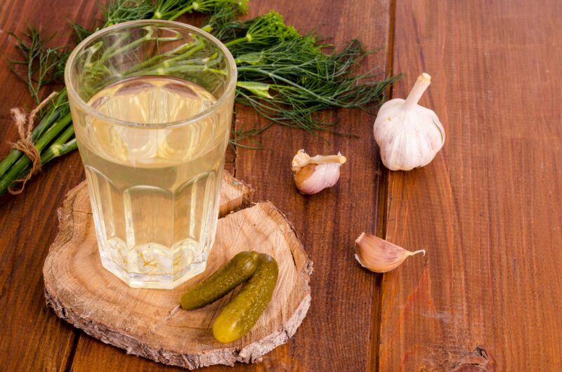 A large glass of pickle juice, next to dill, garlic, and pickles