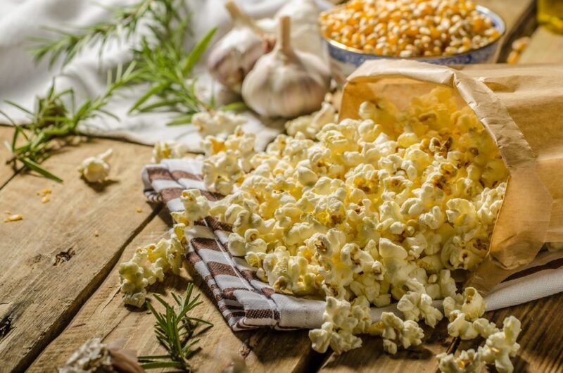 A wooden table with a plate of herbed popcorn, with some herbs and garlic in the background