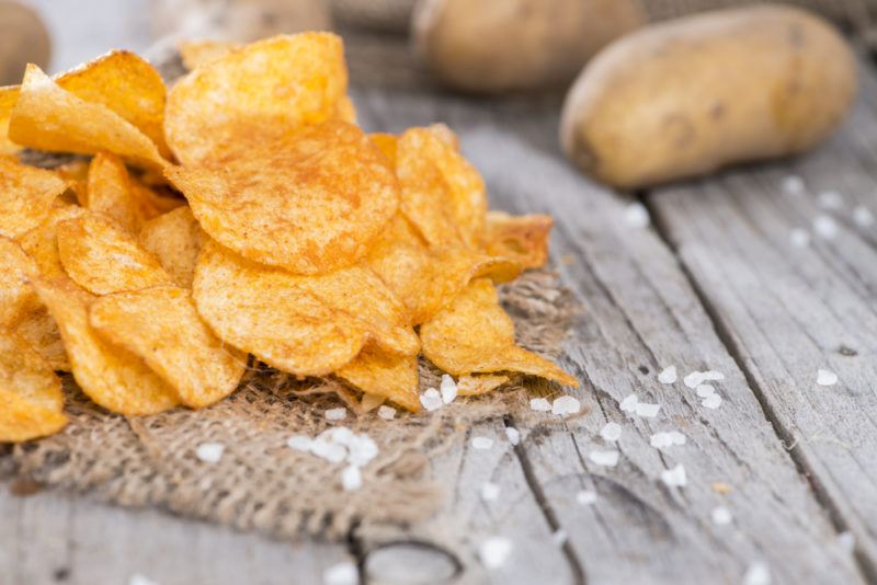 Potato chips with salt on a wooden table with some potatoes in the background