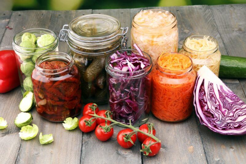 A wooden table with jars of fermented food, plus some ingredients for ferments, like cucumbers, brussels sprouts, and cabbage