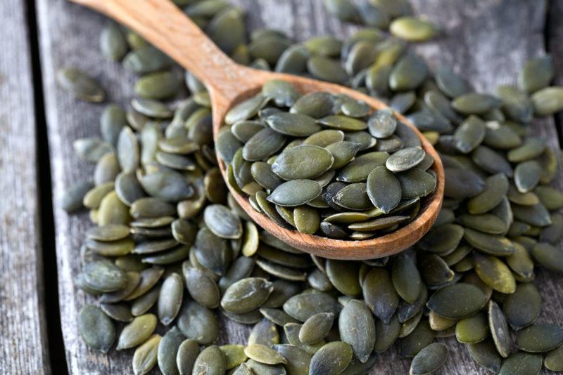 A wooden table with pumpkin seeds scattered across it and more pumpkin seeds on a wooden spoon