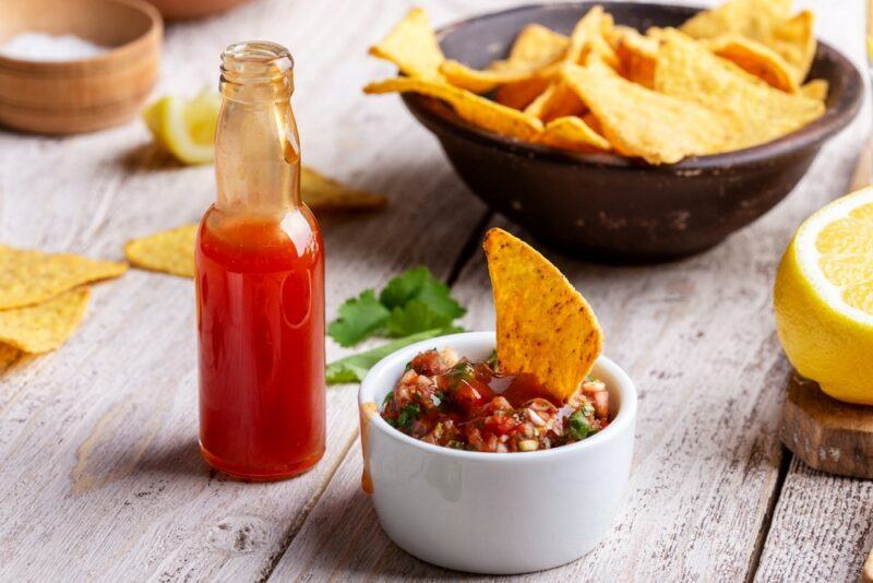 A wooden table with a black bowl of tortilla chips, a small bottle of hot sauce, and a white dish containing salsa, hot sauce, and a single tortilla chip