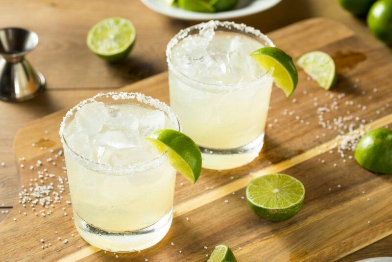 A wooden table with two moonshine margaritas, garnished with a wedge of lime, with limes scattered on the table