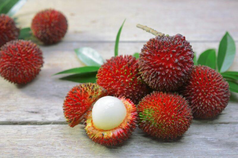 A wooden table, with a collection of red pulasan fruits. One has been opened, showing the white interior