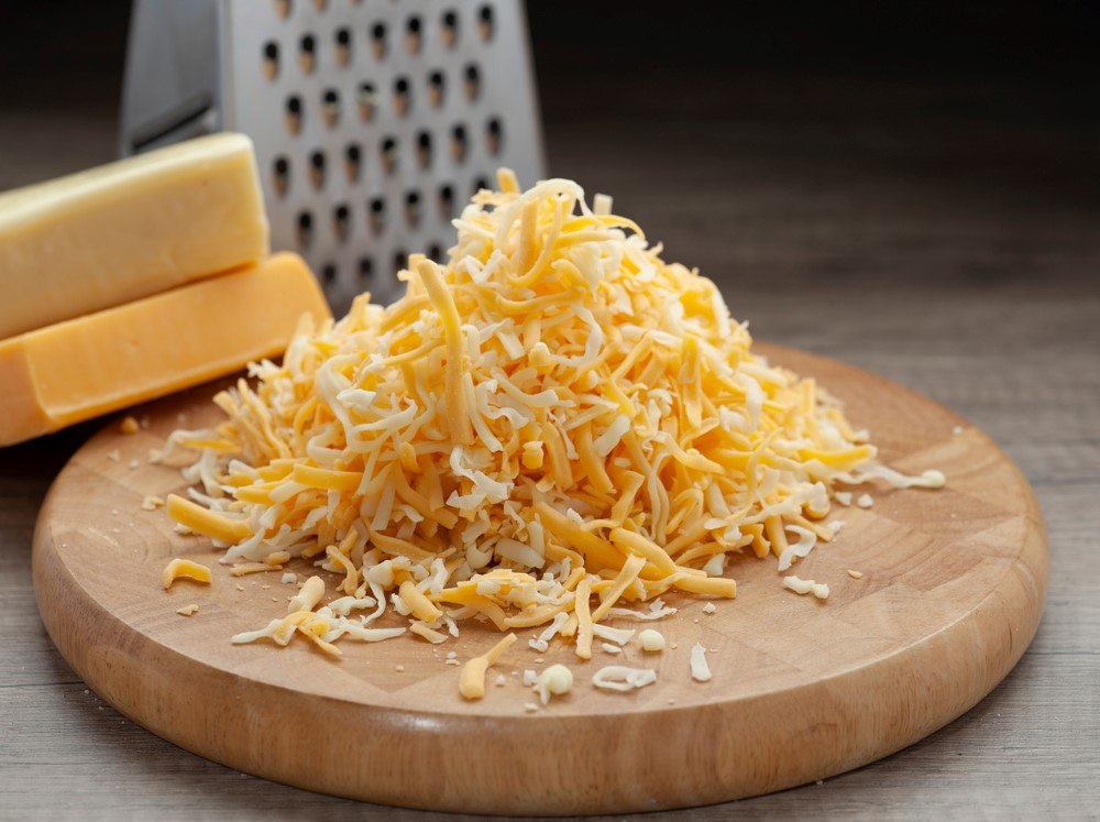 A wooden board with grated cheese in front of a grater and some cheese blocks