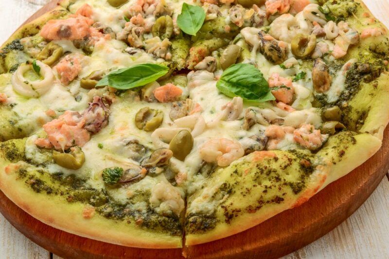 A pizza on a wooden board that uses shrimp, cheese, and pesto as toppings