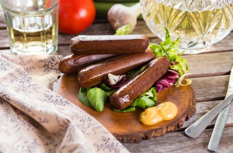 A selection of uncooked plant-based sausages on a wooden board next to a cloth, oil, and a knife and fork