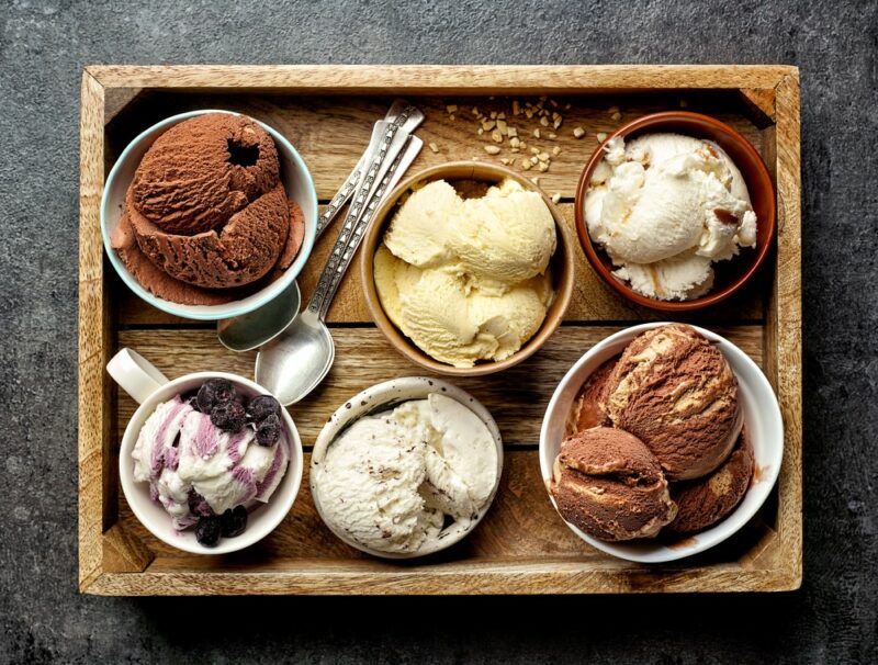 A wooden tray with six mugs and bowls with scoops of different types of ice cream