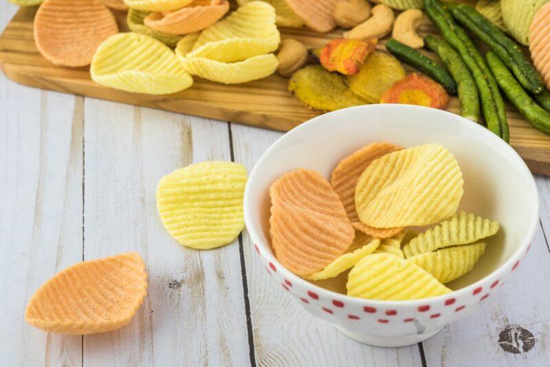 A wooden board with processed veggie chips, a couple of the chips on a table, then a white bowl with more of them