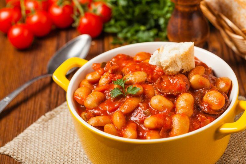 A yellow dish filled with baked beans, with some greens and tomatoes on the table