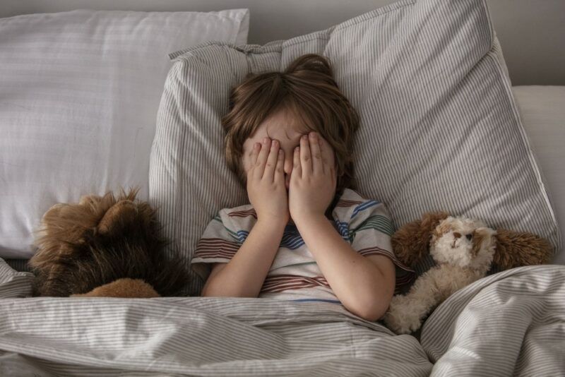 A young boy lying in bed with his hands over his face, next to two stuffed animals. He may have just experienced a nightmare or night terrors.