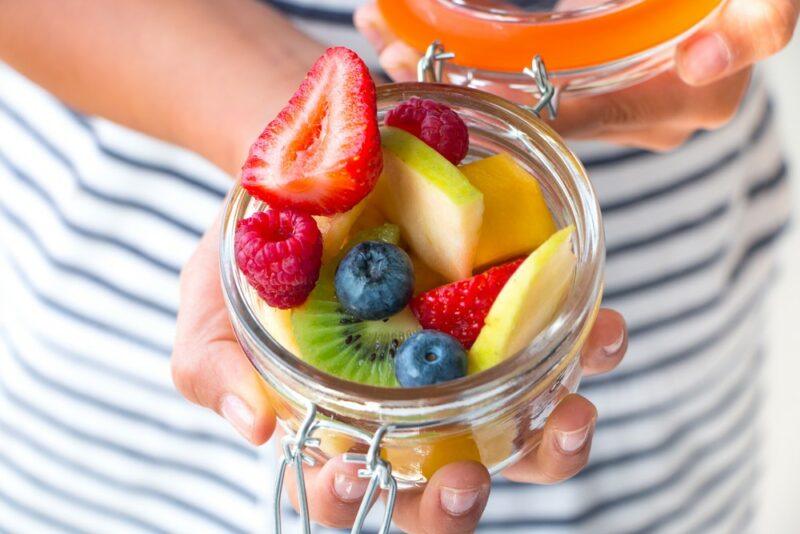 A young child holding a glass jar containing various types of fruit, like apples, blueberries, strawberries, and kiwis