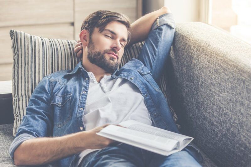 A young man sleeping on a couch in a blue jacket with a book