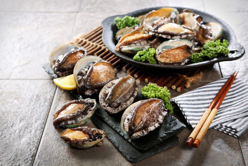 A black dish of ready to eat abalone, with more on the table and some chopsticks