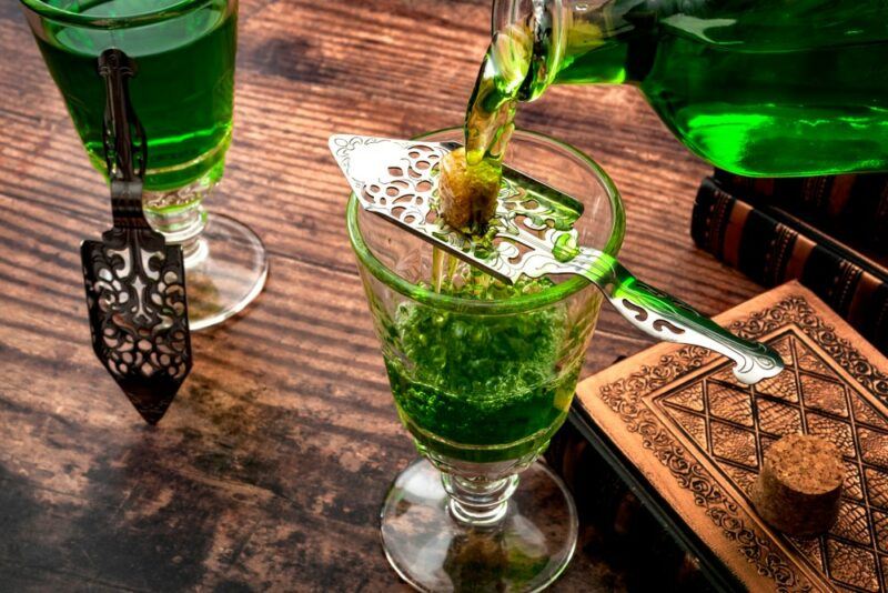 A wooden table with two glasses of absinthe. For one of these, the absinthe is being poured into the glass through a spoon with holes