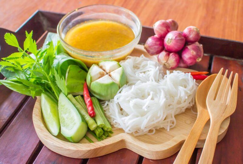 A small wooden board with fresh vegetables, a small glass dish of broth, rice noodles, a fork, and a spoon