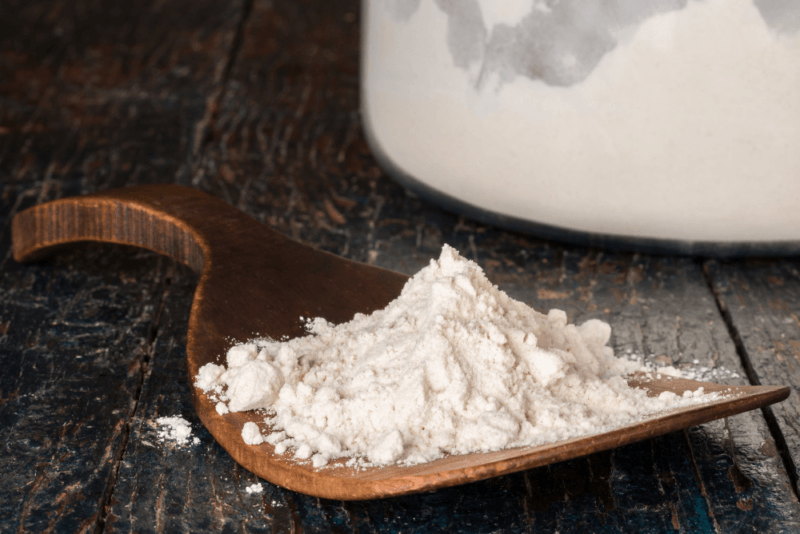 A wooden table with an oddly shaped wooden spoon that contains all purpose flour, plus a jar of all purpose flour in the background