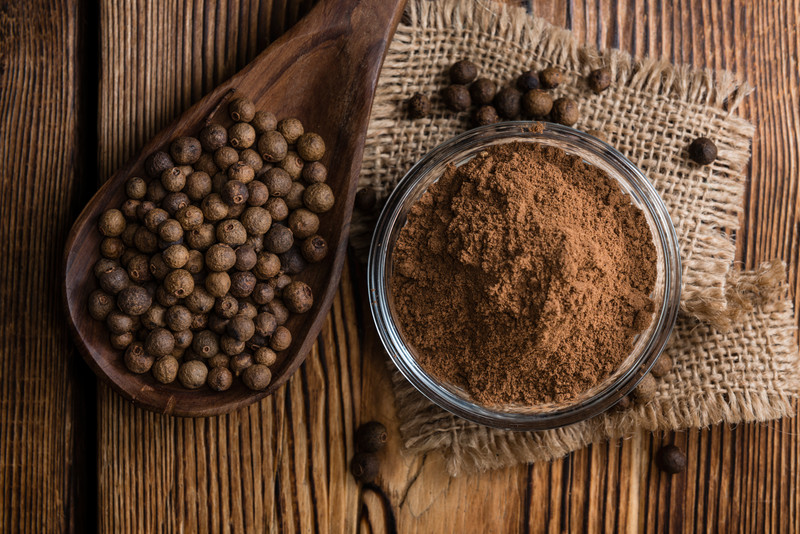 top view image of a small glass bowl on top of a sack cloth full of ground allspice, with wooden ladle beside it full of whole allspice