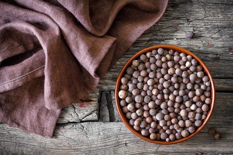 Top view image of allspice in a brown bowl, on a wooden table with red-brown cloth.