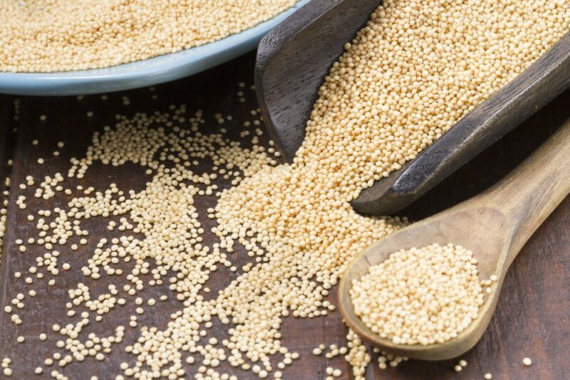 A large scoop of amaranth seeds spilling onto a table, next to a large dish and a wooden spoon also filled with the seeds