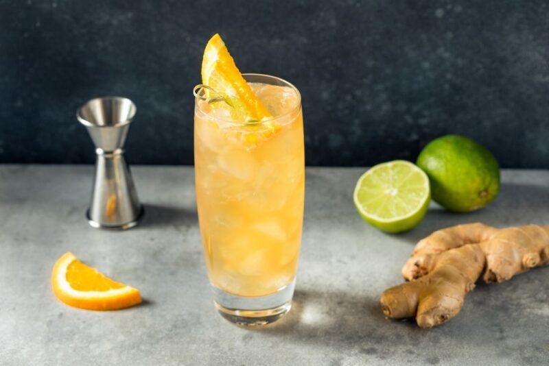 A tall glass containing an anejo highball cocktail with ice, next to a cocktail jigger, a lemon slice, some limes, and ginger