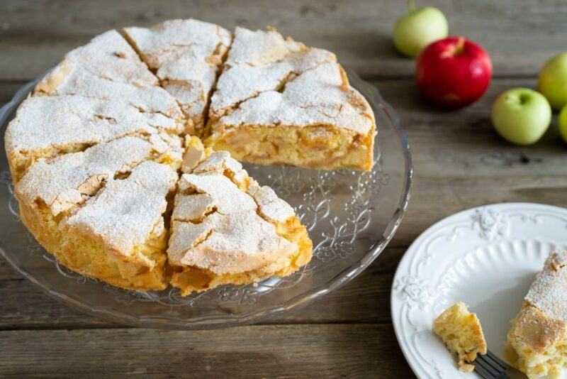 A wooden table with an apple cake that's been cut into pieces, with one piece cut out entirely and put on a different plate. There are also some green apples and one red apple in the background