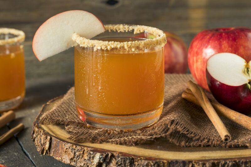 A glass containing an apple pie cocktail, with cinnamon sugar and apple on the rim, next to a second glass, cinnamon sticks, and apple pieces