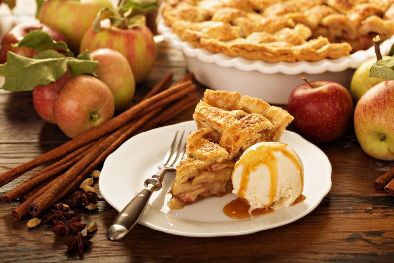 A slice of apple pie on a plate with a fork and a scoop of vanilla ice cream, in front of a large apple pie