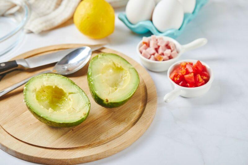 A white table with a board, an avocado that has been cut in half and some fillings. 