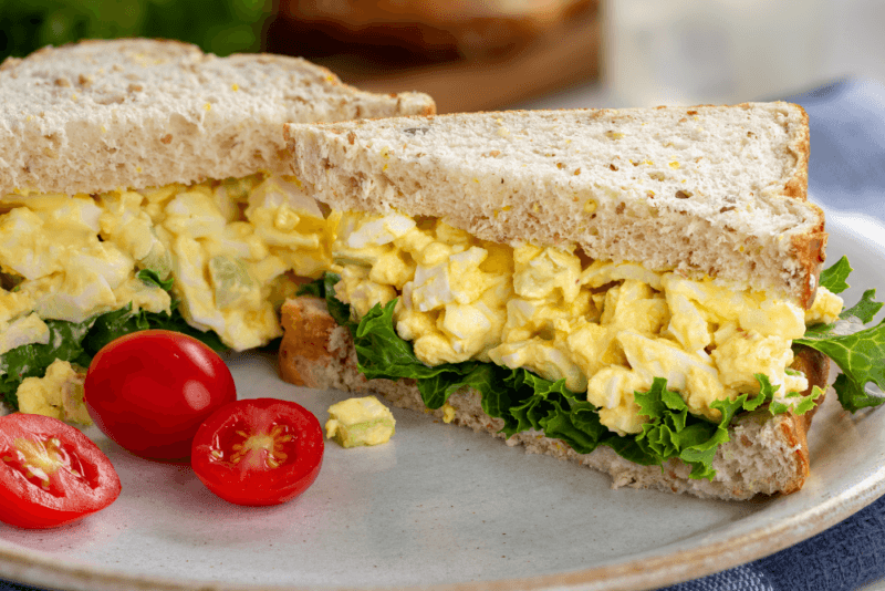 Two halves of an egg salad sandwich with lettuce on the base, plus a few cherry tomato halves on the plate. 