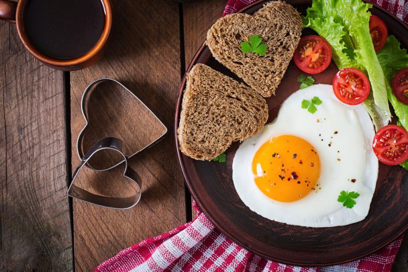 A brown plate with a love heart egg, two pieces of love heart toast, and veggies, on a wooden table next to the heart cutters used