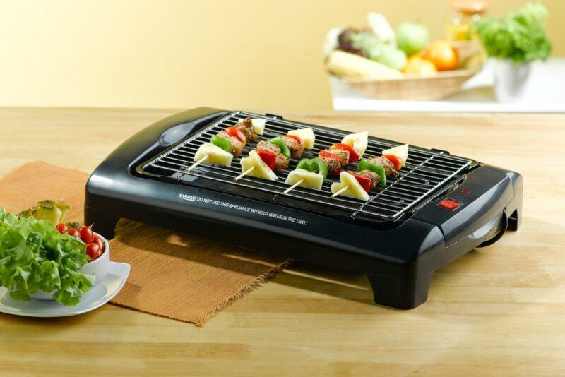 A black indoor grill on a kitchen counter with four kebabs, with containers of vegetables in the foreground and background