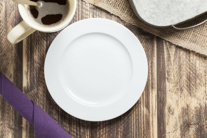 A wooden table with a white empty plate next to a mug of hot coffee