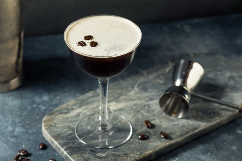 A glass containing an espresso martini with three espresso beans as a garnish, next to a cocktail jigger