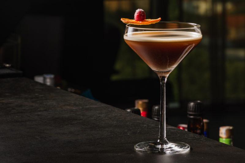 An espresso martini with a raspberry and orange as a garnish on a black bar