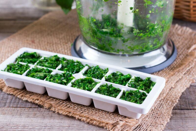 A white ice cube tray filled with crushed herbs and oil, with a blender in the background