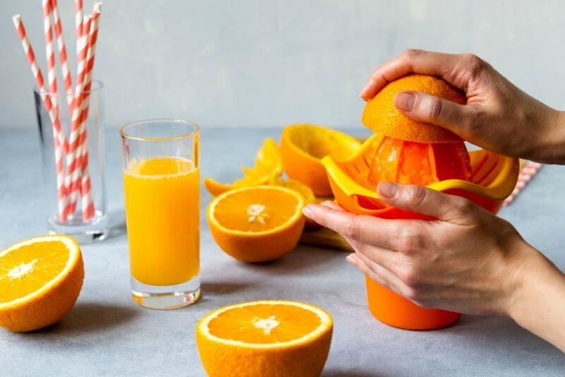 Orange juice being squeezed from a juicer by a pair of hands, with a glass of orange juice on the table and various orange halves