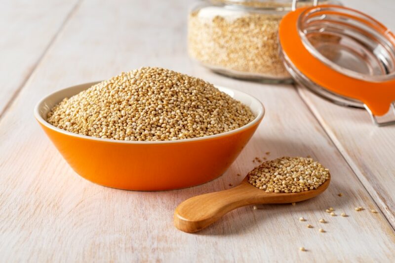 An orange bowl filled with quinoa, next to a spoon of quinoa, with more in a jar in the background