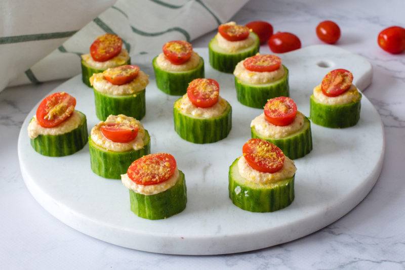 A marble cutting board with cucumber rounds that each have hummus and half a cherry tomato to make appetizers