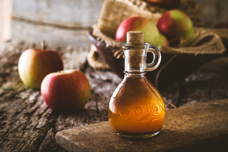 This photo shows a glass flask of apple cider vinegar on a cutting board near two loose apples and a bowl of apples on a wooden table.