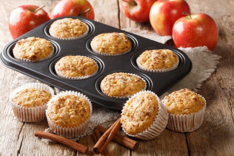 Muffin tins containing apple cinnamon muffins, with more muffins next to the tins, plus cinnamon and apples