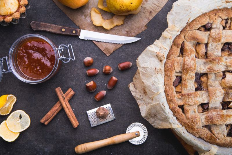 An apple, pear, and hazlenut pie on a black table with nuts and spices next to it