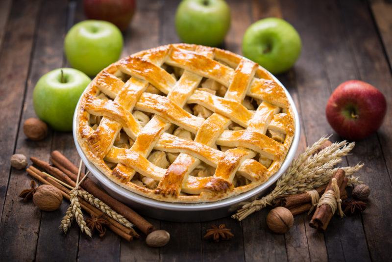 An apple pie on a table next to four green apples and a red apple, along with some spices